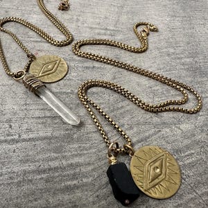 May include: Two gold-toned necklaces with different pendants. One features a clear crystal point wrapped in wire, and a circular charm with an eye design. The other has a black stone and a similar charm. The chains are laid out on a gray surface.