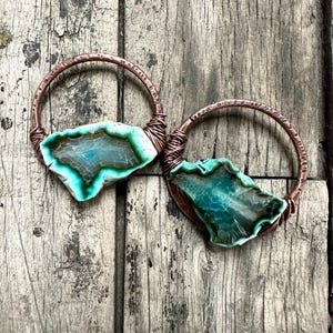 May include: A pair of hoop earrings featuring hammered copper wire frames wrapped around irregular, translucent teal and green agate slices. The stones display natural banding and crystalline textures, secured to the circular metal hoops.