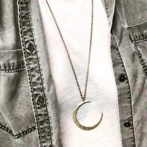 May include: A gold crescent moon pendant necklace on a silver chain. The necklace is worn by a person wearing a white t-shirt and a denim shirt.