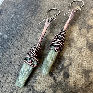 May include: A pair of dangle earrings featuring raw, pale green crystal points wrapped in dark, oxidized copper wire. Each earring has a hammered copper stem with a small, embedded reddish-brown bead and a curved copper ear wire hook.
