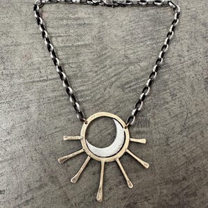 May include: A silver-toned necklace featuring a sun and moon pendant. The pendant has a gold-colored outer ring with silver rays and a crescent moon. The chain is a dark silver color.