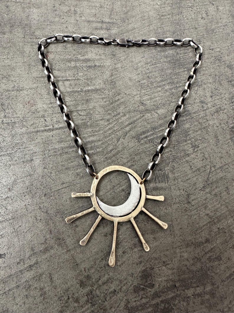 May include: A silver-toned necklace featuring a sun and moon pendant. The pendant has a gold-colored outer ring with silver rays and a crescent moon. The chain is a dark silver color.