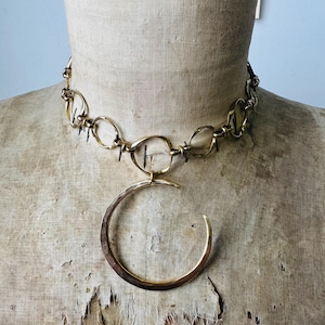 May include: A gold-toned necklace with a large, open circular pendant. The necklace features a chain of interconnected oval links and small spikes. The jewelry is displayed on a vintage mannequin.
