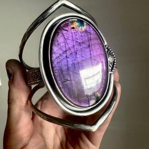May include: A silver cuff bracelet featuring a large, oval-shaped purple labradorite gemstone as the centerpiece. The metal band has a pointed, angular design with intricate engraved floral patterns on the side and a small eye-shaped detail.