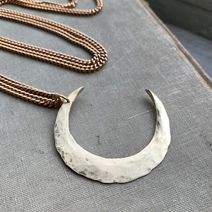 May include: A silver-toned crescent moon pendant necklace with a hammered texture. The pendant hangs from a delicate gold-colored chain. The necklace rests on a gray surface, suggesting a jewelry or accessory theme.