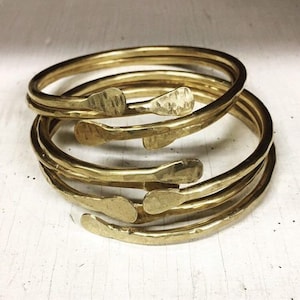 May include: A stack of gold-colored metal bangles. Each bangle has a hammered texture and a unique, flattened end. The bangles are stacked on top of each other, creating a layered effect. The overall design is simple and elegant.