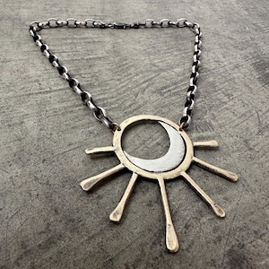 May include: A silver-toned necklace featuring a sun and moon pendant. The pendant has a gold-colored ring with silver rays and a crescent moon. The chain is a silver-toned, chunky link style. The necklace is displayed on a gray surface.