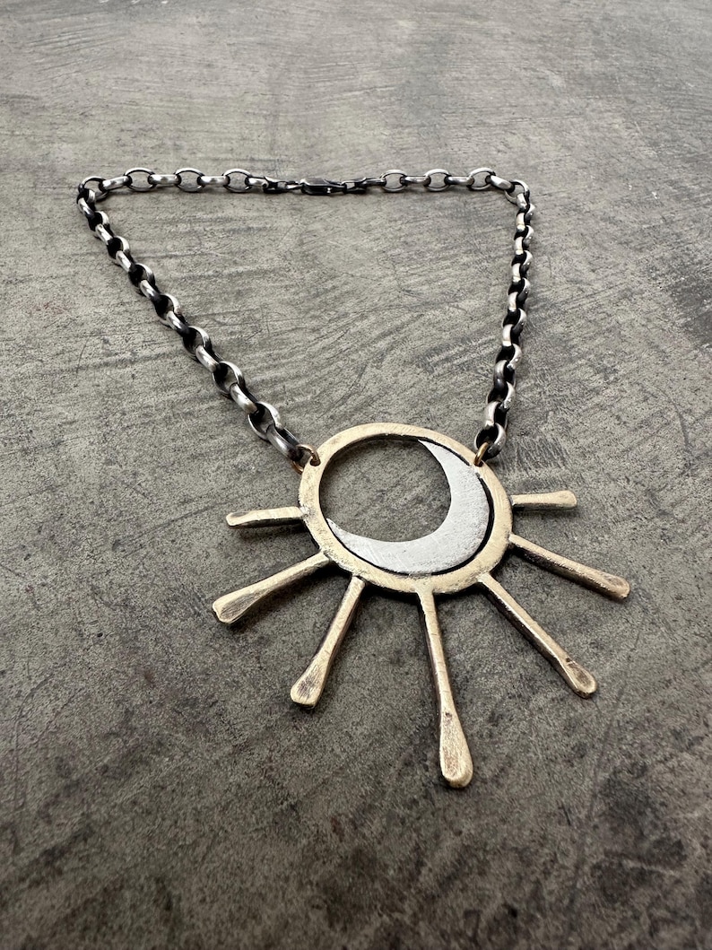May include: A silver-toned necklace featuring a sun and moon pendant. The pendant has a gold-colored ring with silver rays and a crescent moon. The chain is a silver-toned, chunky link style. The necklace is displayed on a gray surface.