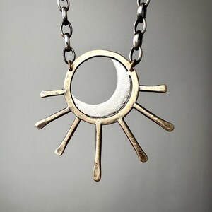 May include: A celestial-themed necklace featuring a silver crescent moon within a gold-toned sunburst pendant. The pendant is suspended from a silver chain. The design is minimalist and geometric, with a focus on the contrast between the silver and gold metals.
