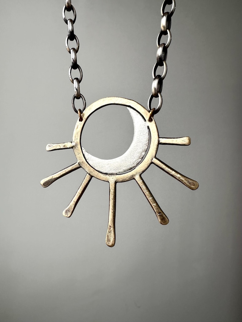May include: A celestial-themed necklace featuring a silver crescent moon within a gold-toned sunburst pendant. The pendant is suspended from a silver chain. The design is minimalist and geometric, with a focus on the contrast between the silver and gold metals.