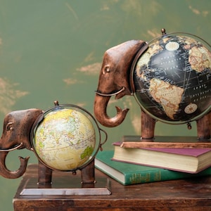 May include: Two decorative metal elephant figurines holding globes. The elephants are brown and the globes are black and white. The globes are on stands and the elephants are on a wooden surface.