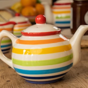 May include: A striped ceramic teapot with a white base and bands of blue, green, yellow, orange, and red. The lid is white with a red knob. The teapot has a curved handle and a spout. Other matching items are visible.