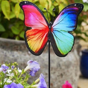 May include: A vibrant metal butterfly garden ornament. The butterfly's wings display a spectrum of colours, including red, orange, yellow, blue, and green. The black stake is set amongst purple flowers and foliage.