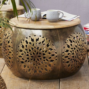May include: A round, gold-toned metal coffee table with intricate cut-out designs. The table has a wooden top and is sitting on a wooden floor.