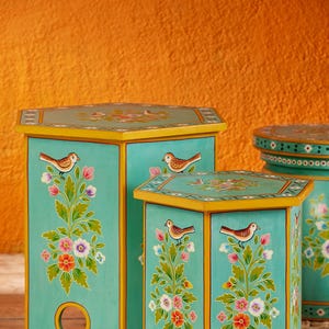 May include: Two turquoise blue hexagonal wooden stools with yellow trim and hand-painted floral designs featuring birds. The stools are on a woven rug.
