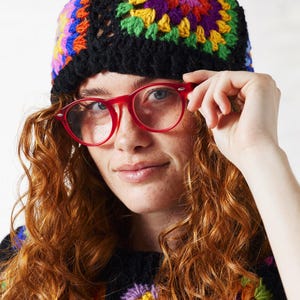 May include: A woman with red curly hair wearing a black crochet hat with colourful granny squares and red round glasses.