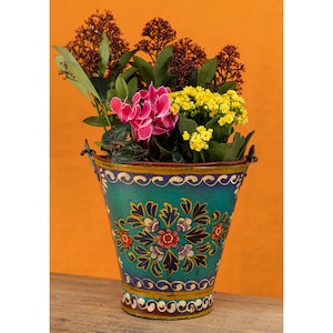 May include: A teal metal bucket planter featuring hand-painted floral motifs in red, blue, and white. The bucket contains a vibrant arrangement of pink cyclamen flowers, yellow kalanchoe blossoms, and dark green foliage with deep red berries.