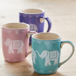 May include: Three ceramic mugs in pink, teal, and purple, each featuring a white elephant design. The mugs have a decorative lace pattern around the rim and a curved handle. They are set on a light wooden surface.