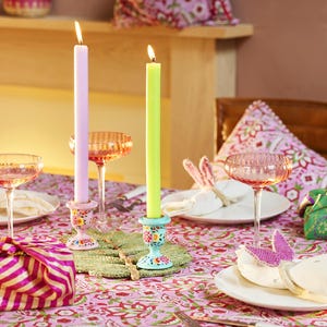 May include: A colourful table setting with a pink floral tablecloth, featuring lit candles in pastel shades, decorative candle holders, and pink champagne glasses. The scene includes wrapped gifts and decorative napkins, creating a celebratory atmosphere.