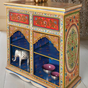 May include: A hand-painted wooden cabinet with intricate floral designs in shades of red, yellow, blue, and green. The cabinet has two drawers and two open shelves, and features a silver elephant figurine and two bowls.