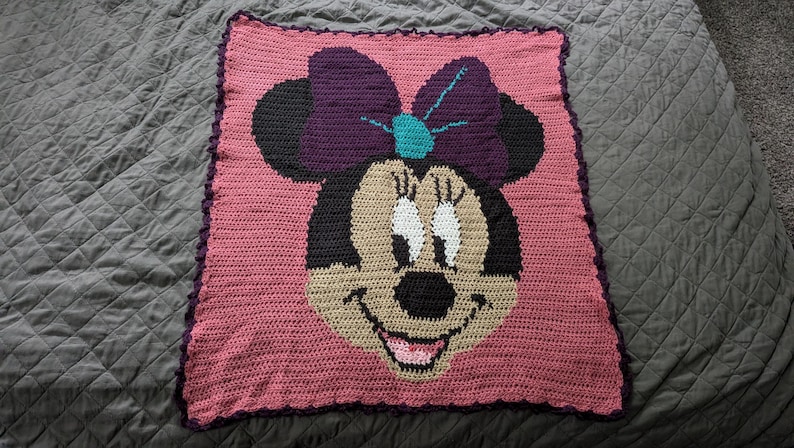 May include: A handmade crochet blanket featuring Minnie Mouse's face. The blanket has a pink background with a black, tan, purple, and teal Minnie Mouse design. The blanket is square-shaped with a dark purple border.