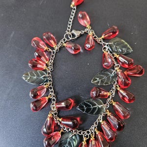 May include: A silver bracelet with red teardrop beads and green leaf charms. The bracelet has a chain design and a clasp. The translucent red beads are attached to the chain with small gold-toned connectors.