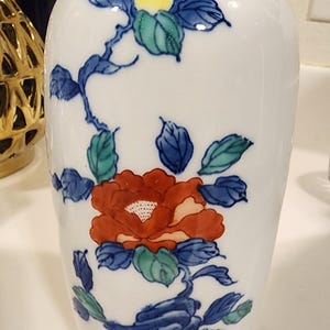 Puede incluir: Jarrón de cerámica blanca con diseño floral. El jarrón presenta una flor roja con hojas y tallos azules y verdes. Hay detalles amarillos y verdes adicionales. El jarrón tiene un cuerpo redondeado y un cuello estrecho.