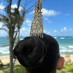 Puede incluir: Un peine decorativo para el cabello con un patrón de carey se inserta en un moño. El peine presenta un diseño vertical adornado con recortes geométricos y una parte superior en forma de estrella. El fondo muestra una playa y cielo azul.