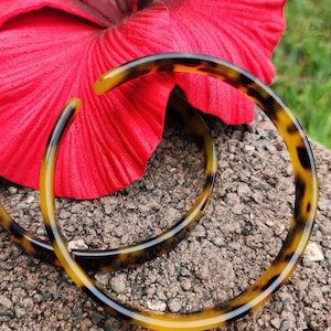Puede incluir: Dos grandes aros de pendientes de carey marrón y amarillo. Los pendientes son gruesos y tienen un acabado brillante. Los pendientes están fotografiados sobre una superficie rocosa marrón con una flor de hibisco roja detrás.