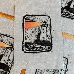 May include: A gray fabric square with a black and orange lighthouse print. The lighthouse is on a rocky shore, with a bright orange light emanating from the tower. The print is framed in a black rectangle.