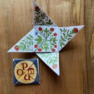 May include: A star-shaped paper craft with a floral design in red, green, and white, alongside a small square box with the letters "P O U" in a circle. The items are on a wooden surface.