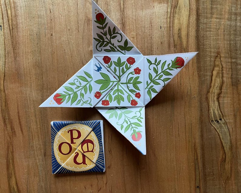 May include: A star-shaped paper craft with a floral design in red, green, and white, alongside a small square box with the letters "P O U" in a circle. The items are on a wooden surface.