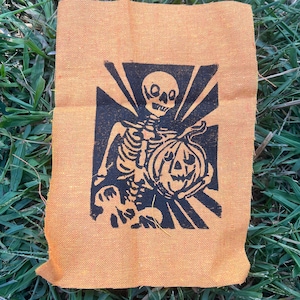 Könnte beinhalten: Ein rechteckiges Stück orangefarbener Stoff mit einem schwarzen Siebdruck-Motiv eines Skeletts, das einen Kürbis hält. Das Skelett ist vor einem Hintergrund aus strahlenden schwarzen Linien positioniert, was einen starken Kontrast auf dem strukturierten Stoff erzeugt.