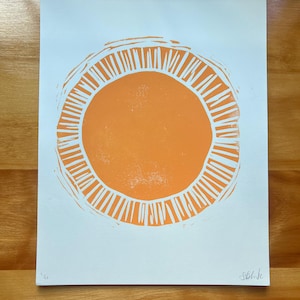 May include: Orange sun linocut print on white paper. The sun design features a solid orange center with a ring of vertical lines around it. The print is signed in the bottom right corner and numbered in the bottom left corner.