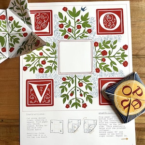 May include: A white paper craft kit with red floral designs, instructions, and a completed star-shaped ornament. The design includes the letters V and O, and the word "LOVE" is visible on a separate piece.