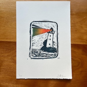 May include: A monochrome linocut print of a lighthouse with a red and orange beam. The lighthouse stands on a rocky outcrop, with waves depicted below. The print is on white paper and signed by the artist.