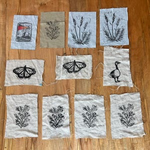 Pode incluir: Uma coleção de quadrados de tecido com vários desenhos de impressão em bloco a preto e branco. As estampas incluem um farol, flores, trigo, borboletas e um ganso. O tecido tem uma borda crua e inacabada.
