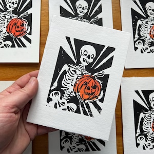 May include: Black and white linocut print featuring a skeleton holding an orange jack-o'-lantern. The artwork has a Halloween theme, with a bold, graphic style. Several prints are visible, suggesting a series or collection.