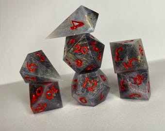 May include: A set of seven polyhedral dice in various shapes. The dice are a smoky gray color with red numbering and gold accents. The dice are arranged in a balanced stack.