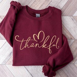 May include: A maroon sweatshirt with the word "thankful" in a gold script font. The word "thankful" has a heart shape above the "k". The sweatshirt is folded on a light brown and white wooden surface.