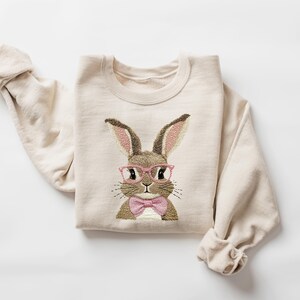 K&ouml;nnte beinhalten: Cremefarbenes Sweatshirt mit einem gestickten Hasen mit rosa Brille und rosa Fliege. Der Hase hat braunes und hellbraunes Fell mit rosa Innenohren. Die &Auml;rmel sind hochgekrempelt, auf wei&szlig;em Hintergrund.