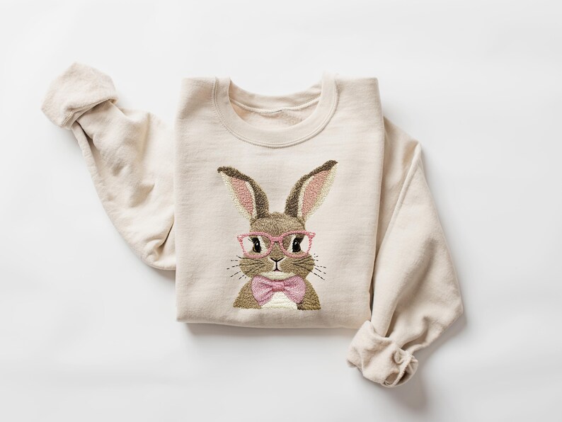 K&ouml;nnte beinhalten: Cremefarbenes Sweatshirt mit einem gestickten Hasen mit rosa Brille und rosa Fliege. Der Hase hat braunes und hellbraunes Fell mit rosa Innenohren. Die &Auml;rmel sind hochgekrempelt, auf wei&szlig;em Hintergrund.