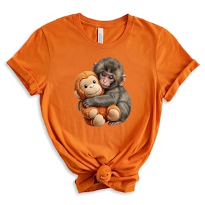 May include: Orange t-shirt featuring a graphic of two monkeys hugging. One monkey is gray and the other is a plush toy in shades of brown and tan. The t-shirt is made of a soft material.