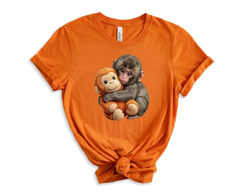 May include: Orange t-shirt featuring a graphic of two monkeys hugging. One monkey is gray and the other is a plush toy in shades of brown and tan. The t-shirt is made of a soft material.