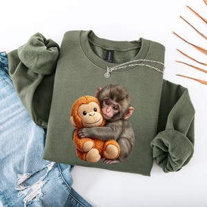 May include: An olive green sweatshirt featuring a graphic of a monkey hugging a stuffed monkey. The sweatshirt is paired with light blue denim jeans. A silver necklace is draped on the sweatshirt. The image is set against a white background.