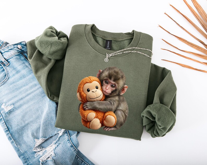 May include: An olive green sweatshirt featuring a graphic of a monkey hugging a stuffed monkey. The sweatshirt is paired with light blue denim jeans. A silver necklace is draped on the sweatshirt. The image is set against a white background.