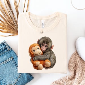 May include: Cream-colored t-shirt featuring a graphic of a gray monkey hugging an orange stuffed monkey. The t-shirt is folded and surrounded by blue jeans, white shoes, and a beige scarf.