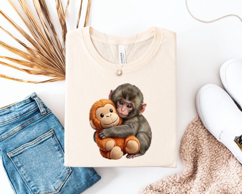May include: Cream-colored t-shirt featuring a graphic of a gray monkey hugging an orange stuffed monkey. The t-shirt is folded and surrounded by blue jeans, white shoes, and a beige scarf.