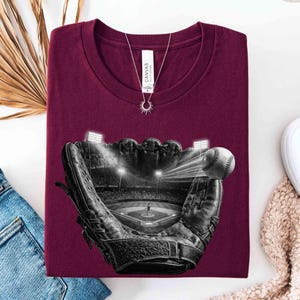 May include: Burgundy t-shirt featuring a baseball glove with a baseball and a baseball field scene inside. The shirt has a crew neck and a silver necklace. The shirt is folded on a white surface.