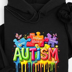 May include: Black hoodie with a colorful graphic design. The design features puzzle pieces with the words "Always", "Unique", "Intelligent", "Always", and "Sometimes". The word "AUTISM" is in large, colorful letters with a dripping paint effect.